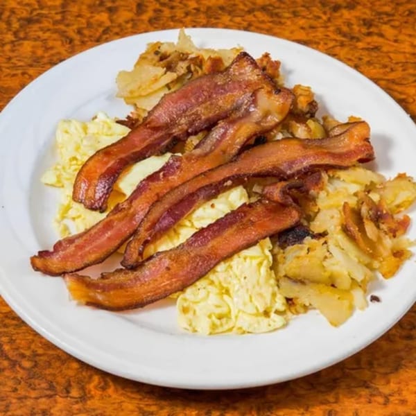 A diner breakfast plate with crispy bacon strips, scrambled eggs, and home fries on a white plate.