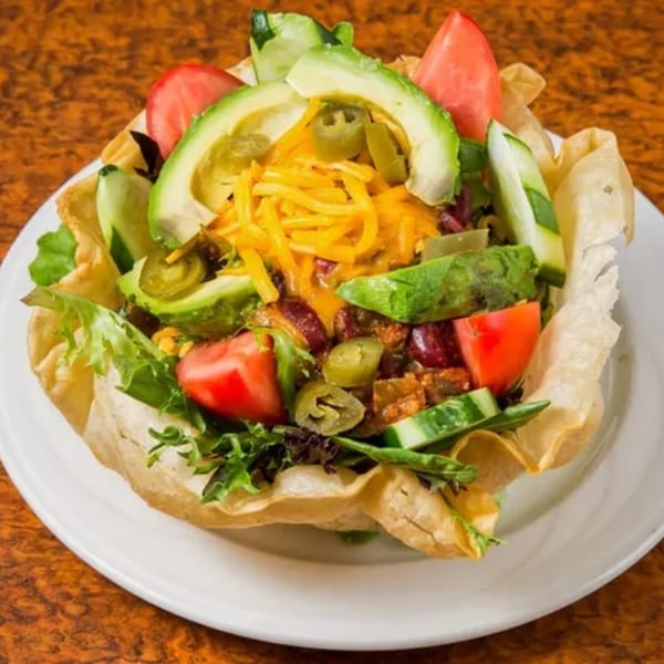 A taco salad in a crispy tortilla bowl with lettuce, tomatoes, avocado, cheese, and jalapeños.