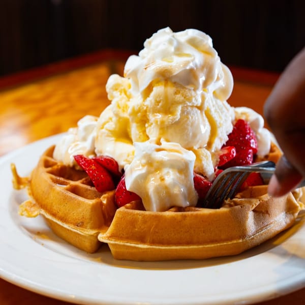 A hand with a fork cutting into a waffle topped with strawberries, whipped cream, and ice cream.