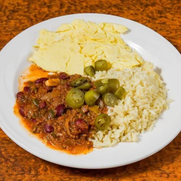 A diner plate with chili, rice, scrambled eggs, and sliced jalapeños on a brown table.