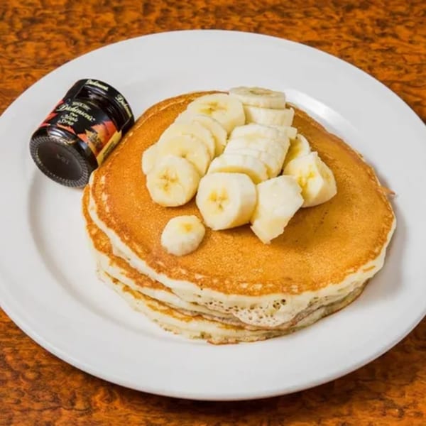 Stack of golden pancakes topped with sliced bananas and a small bottle of syrup on a white plate.